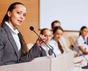 une jeune femme devant un pupitre et un micro qui prend la parole en public sans stress