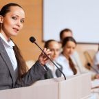 une jeune femme devant un pupitre et un micro qui prend la parole en public sans stress